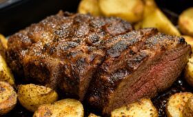 Fotografia de primeiro plano de uma peça suculenta de carne assada fatiada, cercada por batatas douradas e temperadas no forno, servida em uma assadeira preta.