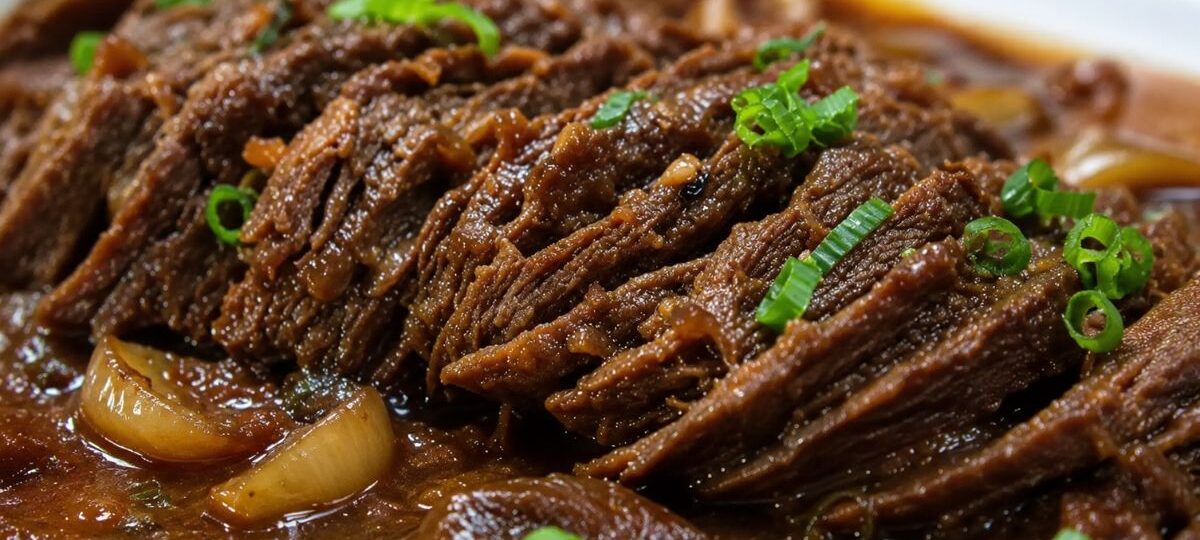Uma foto em close detalha fatias de carne bovina assada, desfiando levemente, submersas em um molho escuro e brilhante. Pedaços de cebola caramelizada estão visíveis no molho e o prato está finalizado com cebolinha verde picada salpicada por cima.