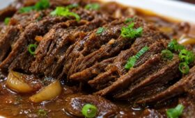 Uma foto em close detalha fatias de carne bovina assada, desfiando levemente, submersas em um molho escuro e brilhante. Pedaços de cebola caramelizada estão visíveis no molho e o prato está finalizado com cebolinha verde picada salpicada por cima.