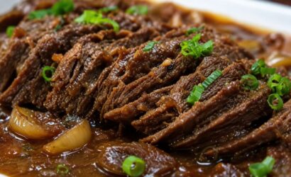 Uma foto em close detalha fatias de carne bovina assada, desfiando levemente, submersas em um molho escuro e brilhante. Pedaços de cebola caramelizada estão visíveis no molho e o prato está finalizado com cebolinha verde picada salpicada por cima.