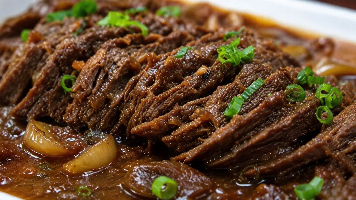 Uma foto em close detalha fatias de carne bovina assada, desfiando levemente, submersas em um molho escuro e brilhante. Pedaços de cebola caramelizada estão visíveis no molho e o prato está finalizado com cebolinha verde picada salpicada por cima.