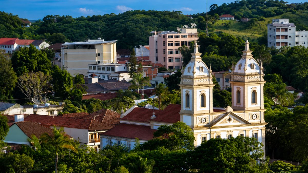 Fotografia aérea do centro de Valença, exibindo em destaque as duas torres brancas da Catedral de Nossa Senhora da Glória, cercada por árvores, telhados coloniais, edifícios e colinas verdes.