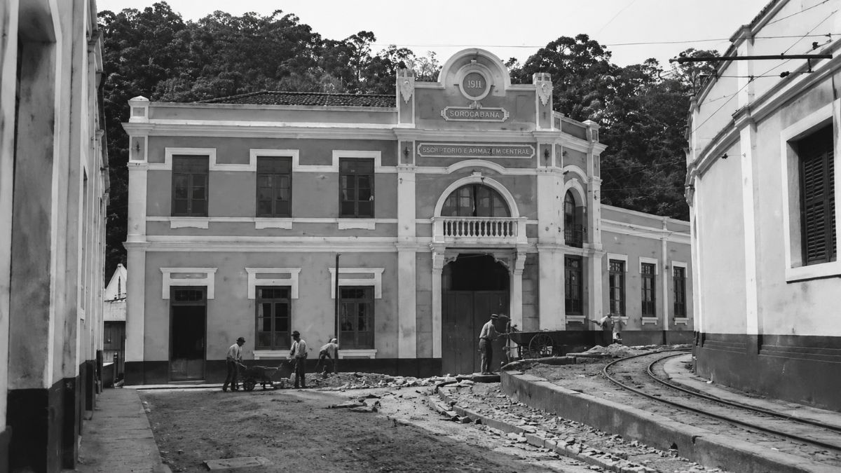 Fotografia antiga em preto e branco mostrando a fachada do edifício da Companhia de Papel Itacolomi em Mendes no Rio de Janeiro com operários trabalhando na rua e trilhos industriais na frente da fábrica.