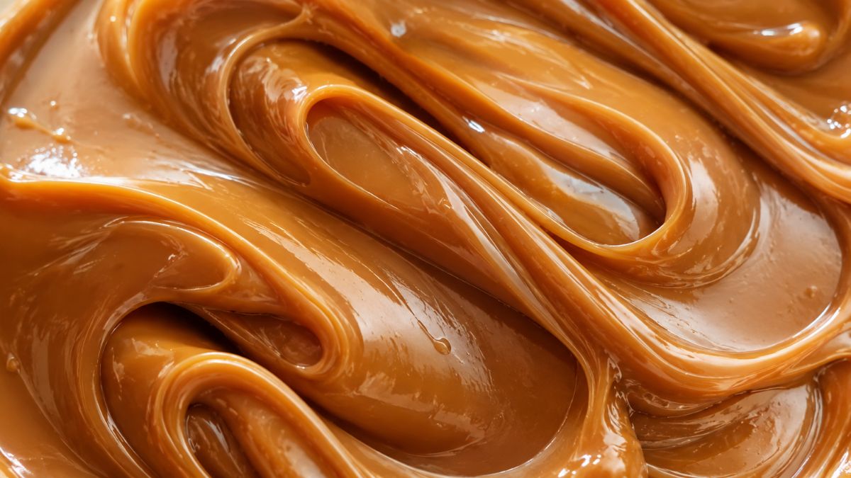 Close-up texturizado de doce de leite caseiro super cremoso e brilhante, mostrando as ondas suaves e a cor caramelo dourado de uma textura aveludada perfeita.