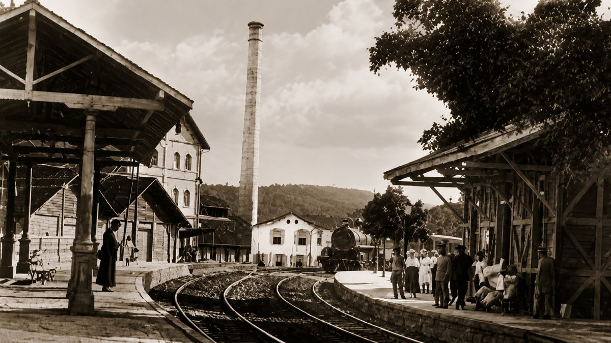 Fotografia antiga em tom sépia mostrando a movimentada Estação Ferroviária Neri Ferreira no município de Mendes, Rio de Janeiro, com passageiros vestidos em trajes de época aguardando nas plataformas cobertas de madeira ao lado dos trilhos e uma antiga locomotiva a vapor posicionada ao centro, com uma grande chaminé industrial e montanhas ao fundo.