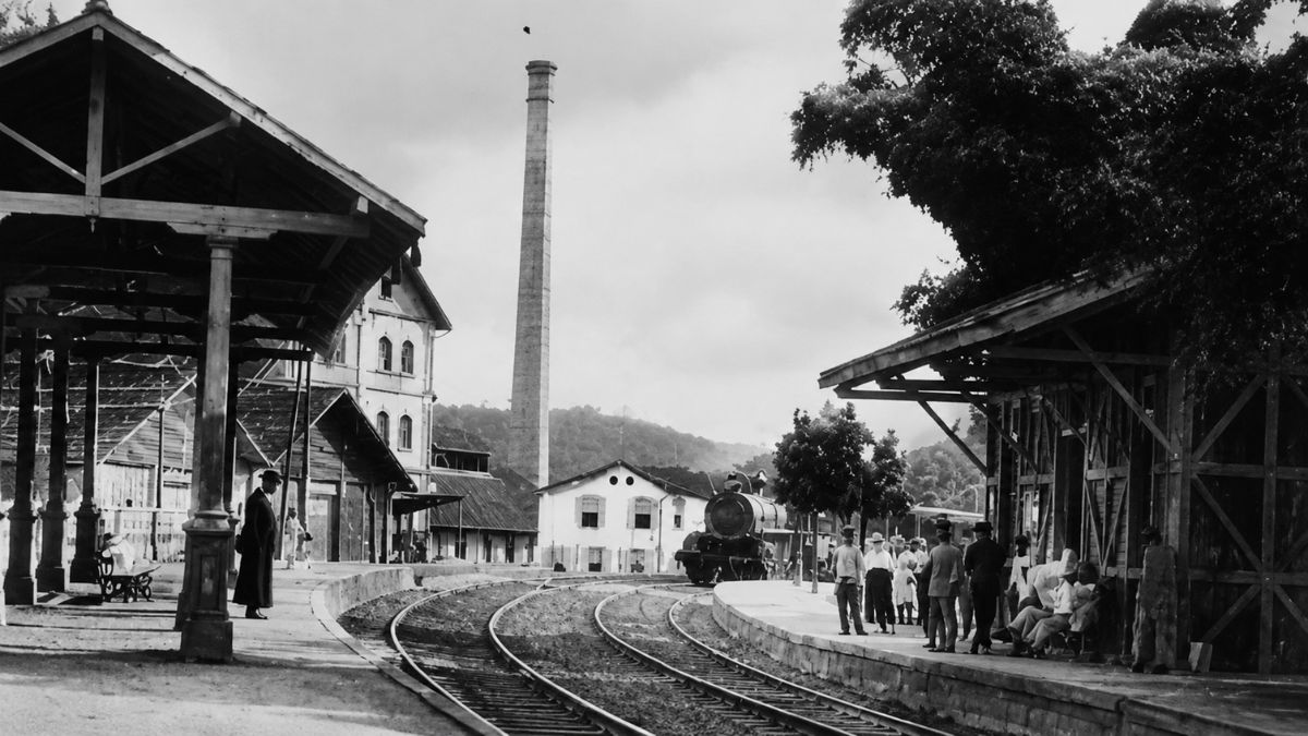 Fotografia antiga em preto e branco da Estação Nery Ferreira em Mendes, Rio de Janeiro, exibindo os trilhos da Estrada de Ferro Dom Pedro II, plataformas de embarque com passageiros e uma chaminé industrial ao fundo no vale do café fluminense.