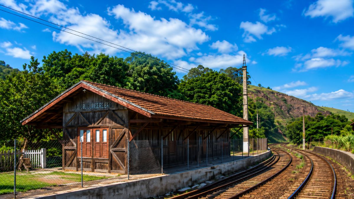 Antiga estação de trem feita de madeira na cidade de Mendes, mostrando a estrutura rústica com telhado de telhas cerâmicas e os trilhos ferroviários em curva sob o céu azul.