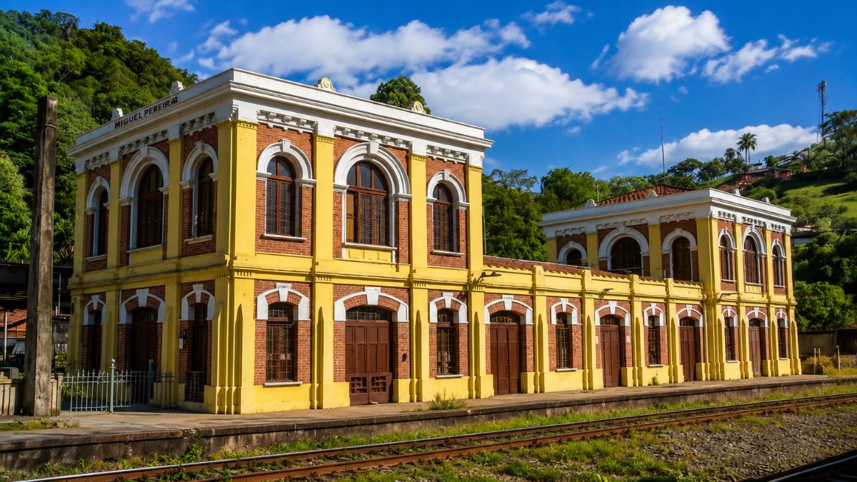 Fachada da antiga estação ferroviária de Engenheiro Paulo de Frontin, exibindo arquitetura colonial com tijolos ecológicos aparentes, janelas em arco e trilhos de trem em primeiro plano.