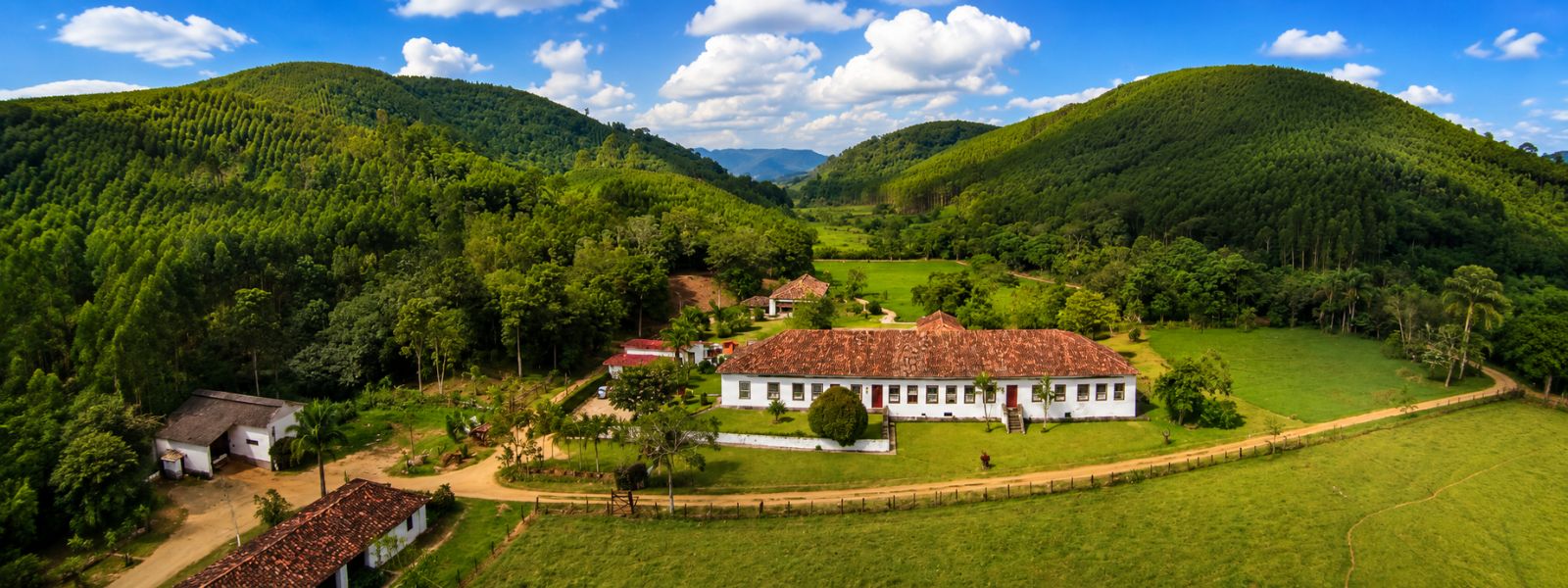 Vista aérea de uma fazenda histórica do Vale do Café com casarão colonial, áreas verdes, estradas rurais e montanhas cobertas por vegetação.