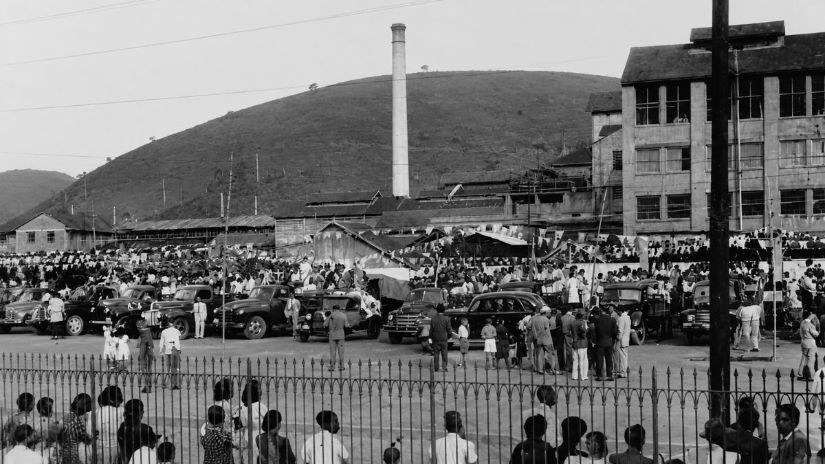 Fotografia antiga em preto e branco de uma grande multidão e diversos carros antigos estacionados para a festa de São Cristóvão em Mendes, em frente a uma fábrica com chaminé.