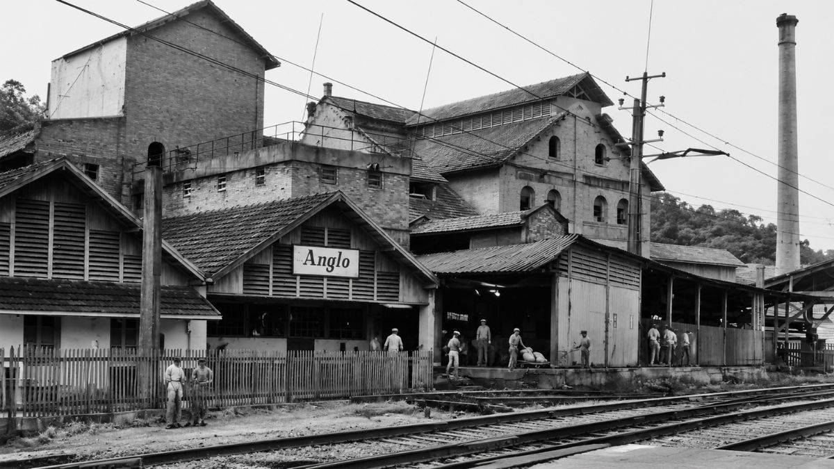 Fotografia antiga em preto e branco do Frigorífico Anglo S/A em Mendes mostrando a fachada com a placa Anglo, operários trabalhando na plataforma de carga e trilhos ferroviários na frente do complexo fabril.