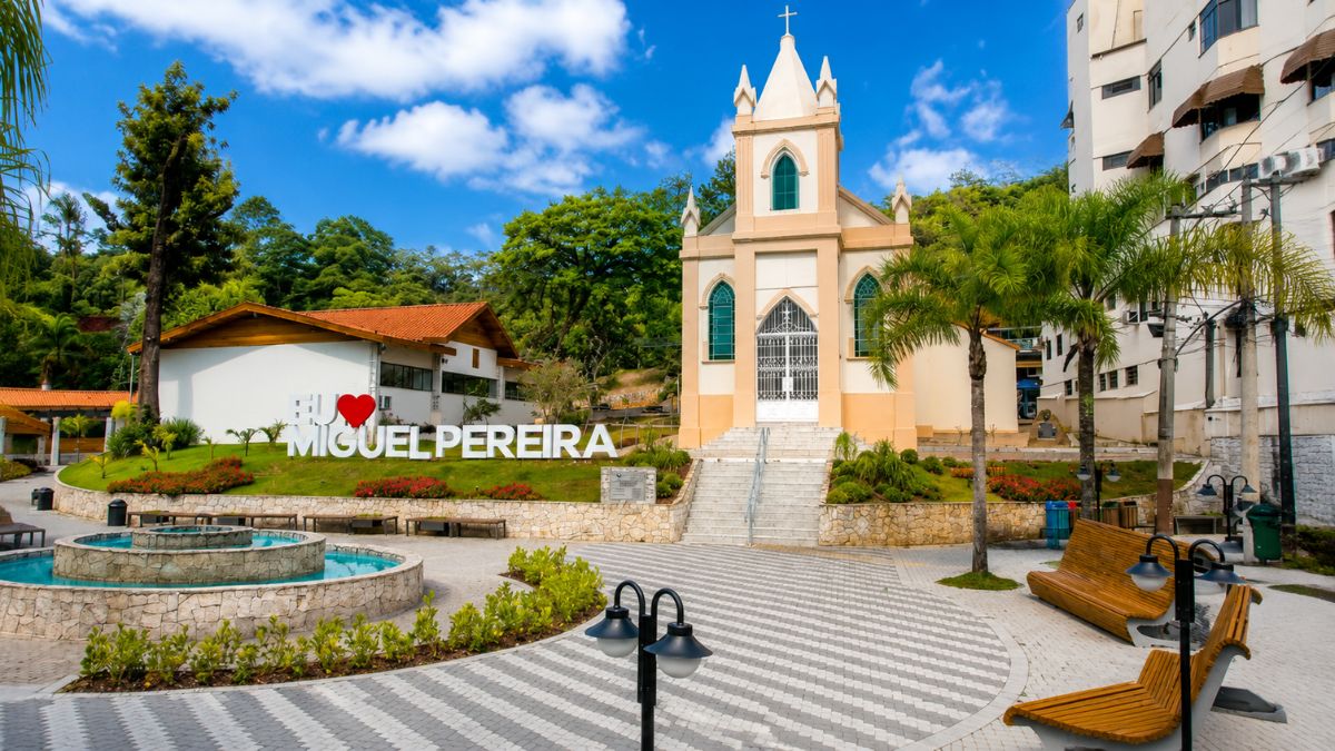 Fotografia da fachada da Igreja Matriz de Miguel Pereira em estilo neoclássico, exibindo a praça central pavimentada com chafariz circular, letreiro turístico e bancos de madeira sob céu azul.