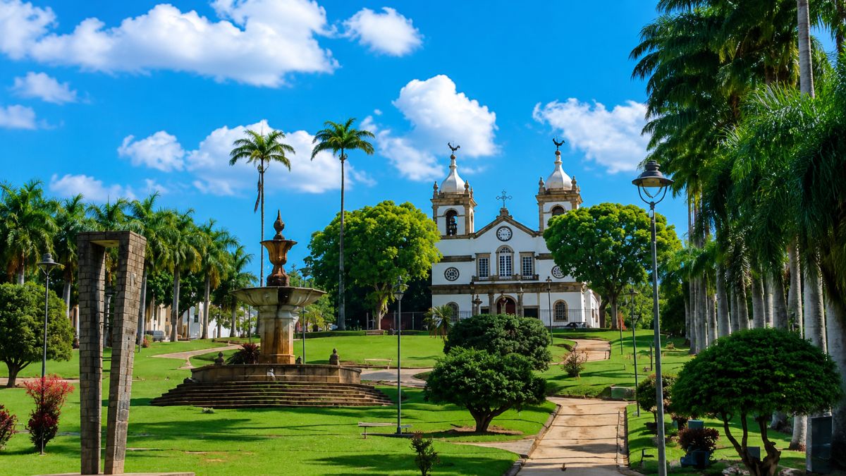 Fotografia da Igreja Matriz de Vassouras ao fundo de uma grande praça com gramado verde, exibindo um chafariz de pedra colonial, palmeiras imperiais altas e caminhos pavimentados sob céu azul.