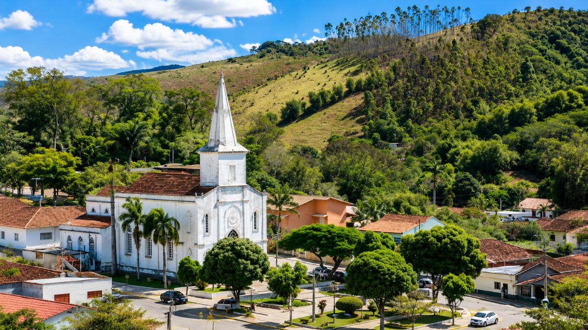 Fotografia aérea da Igreja Matriz de Rio das Flores, exibindo o templo católico branco com torre pontiaguda, praça arborizada, casas coloniais e grandes colinas verdes ao fundo.