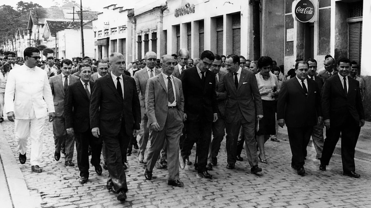 Fotografia antiga em preto e branco mostrando um grupo de homens de terno caminhando em comitiva sobre uma rua de paralelepípedos durante o movimento de emancipação política de Mendes no Rio de Janeiro.