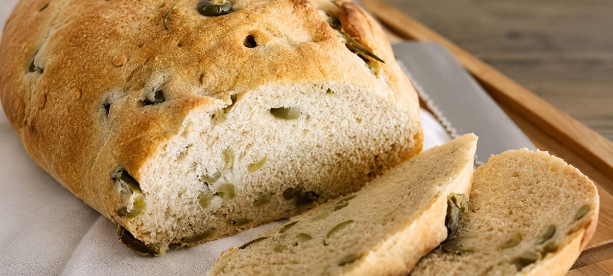 Fotografia em close-up de um pão rústico artesanal dourado, fatiado sobre uma tábua de madeira com pano branco. O pão é recheado e decorado com azeitonas verdes inteiras e ramos de alecrim fresco, mostrando a textura do miolo e das fatias cortadas.