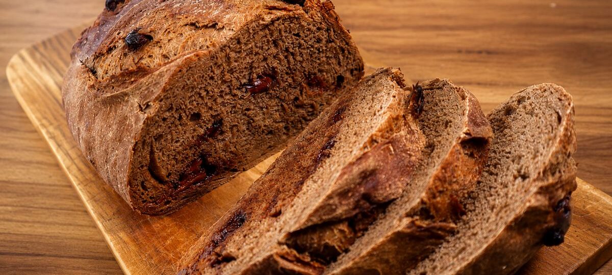 Visão em ângulo alto de um pão de cacau artesanal redondo e rústico, fatiado sobre uma tábua de madeira clara. O pão tem crosta escura e crocante e miolo de cacau macio recheado com pedaços visíveis de frutas vermelhas escuras. Três fatias estão cortadas e empilhadas ao lado do pão principal, revelando o recheio de frutas em cada uma. Fundo de mesa de madeira desfocado.