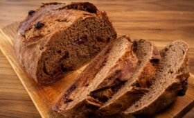 Visão em ângulo alto de um pão de cacau artesanal redondo e rústico, fatiado sobre uma tábua de madeira clara. O pão tem crosta escura e crocante e miolo de cacau macio recheado com pedaços visíveis de frutas vermelhas escuras. Três fatias estão cortadas e empilhadas ao lado do pão principal, revelando o recheio de frutas em cada uma. Fundo de mesa de madeira desfocado.