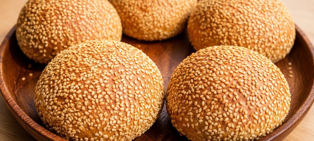 Quatro pães de hambúrguer artesanais dourados e redondos, totalmente cobertos com sementes de gergelim, dispostos em uma bandeja de madeira sobre uma mesa de madeira clara.