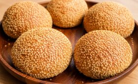 Quatro pães de hambúrguer artesanais dourados e redondos, totalmente cobertos com sementes de gergelim, dispostos em uma bandeja de madeira sobre uma mesa de madeira clara.