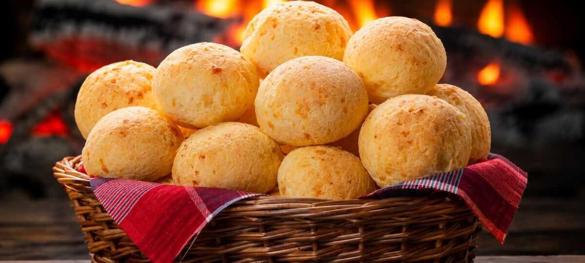 Cesta de vime recheada com pães de queijo da roça dourados sobre mesa de madeira com fogão a lenha aceso ao fundo.