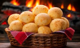 Cesta de vime recheada com pães de queijo da roça dourados sobre mesa de madeira com fogão a lenha aceso ao fundo.
