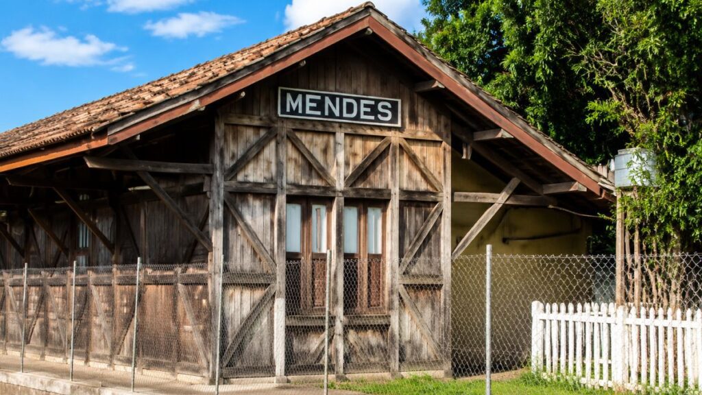 Fotografia colorida em plano médio detalhando a fachada de um antigo galpão de madeira rústica com uma placa preta retangular onde está escrito Mendes em letras brancas garrafais instalada logo abaixo do telhado de telhas de barro. A estrutura de madeira apresenta janelas centrais com molduras de vidro protegidas por uma cerca de arame galvanizado e uma cerca de madeira pintada de branco no canto inferior direito, com vegetação verde e céu azul com nuvens ao fundo.