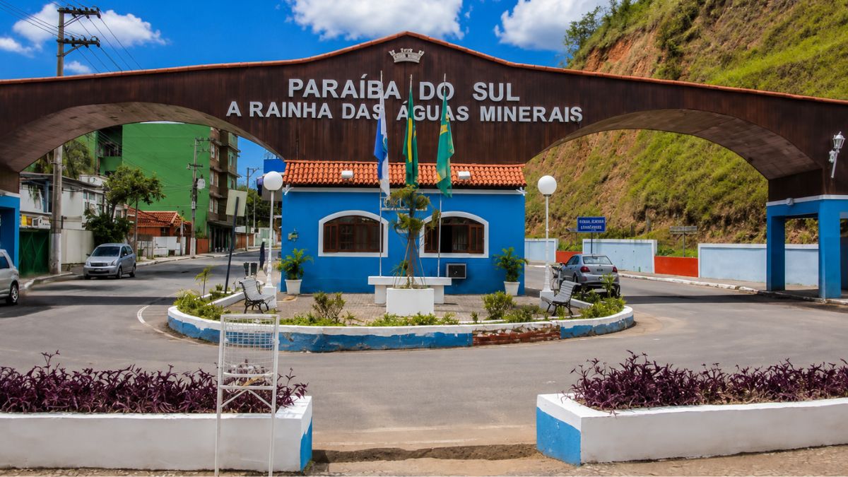 Fotografia do portal de entrada de Paraíba do Sul, exibindo um grande arco de madeira com a inscrição Rainha das Águas Minerais, uma guarita azul com bandeiras e uma rotatória ajardinada.
