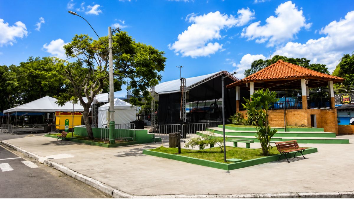 Fotografia da Praça Teixeira Campos em Pinheiral, exibindo um quiosque com telhado colonial, canteiros verdes com bancos de madeira, palcos de eventos e árvores sob céu azul.