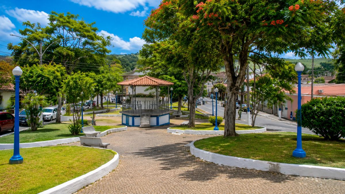 Fotografia da praça central de Rio Claro, exibindo um coreto tradicional com telhado colonial, canteiros gramados com bancos de concreto, postes azuis e árvores floridas sob céu azul.