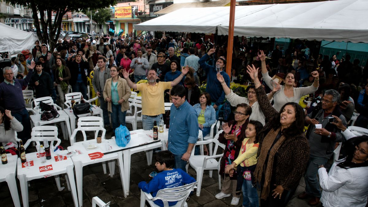 Multidao animada com os bracos levantados e mesas brancas ao ar livre durante evento cultural em Mendes.