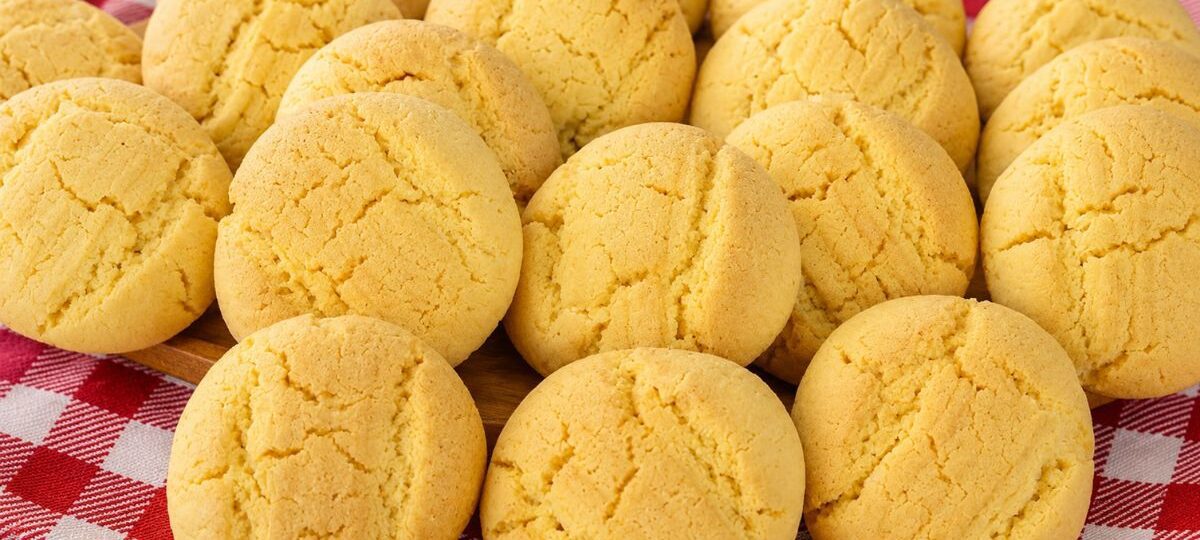 Uma pilha de biscoitos de fubá redondos, amarelos e texturizados, assados e dispostos em cima de um pano de xadrez vermelho e branco sobre uma superfície de madeira.