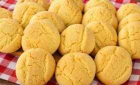 Uma pilha de biscoitos de fubá redondos, amarelos e texturizados, assados e dispostos em cima de um pano de xadrez vermelho e branco sobre uma superfície de madeira.