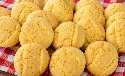 Uma pilha de biscoitos de fubá redondos, amarelos e texturizados, assados e dispostos em cima de um pano de xadrez vermelho e branco sobre uma superfície de madeira.