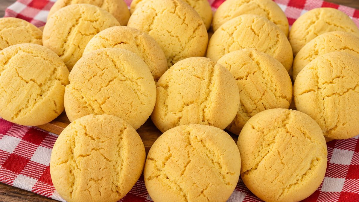 Uma pilha de biscoitos de fubá redondos, amarelos e texturizados, assados e dispostos em cima de um pano de xadrez vermelho e branco sobre uma superfície de madeira.