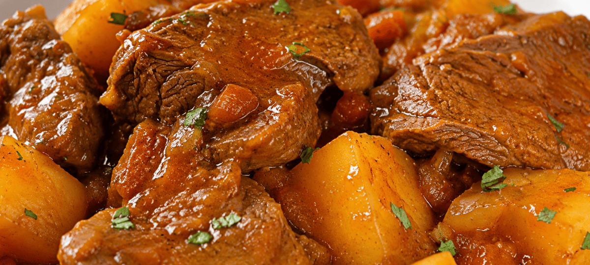 close-up de um prato com fraldinha fatiada em cubos grandes, cozida na panela de pressão, imersa em um molho marrom concentrado com rodelas de cebolas cozidas. O prato está servido com arroz branco e polenta.