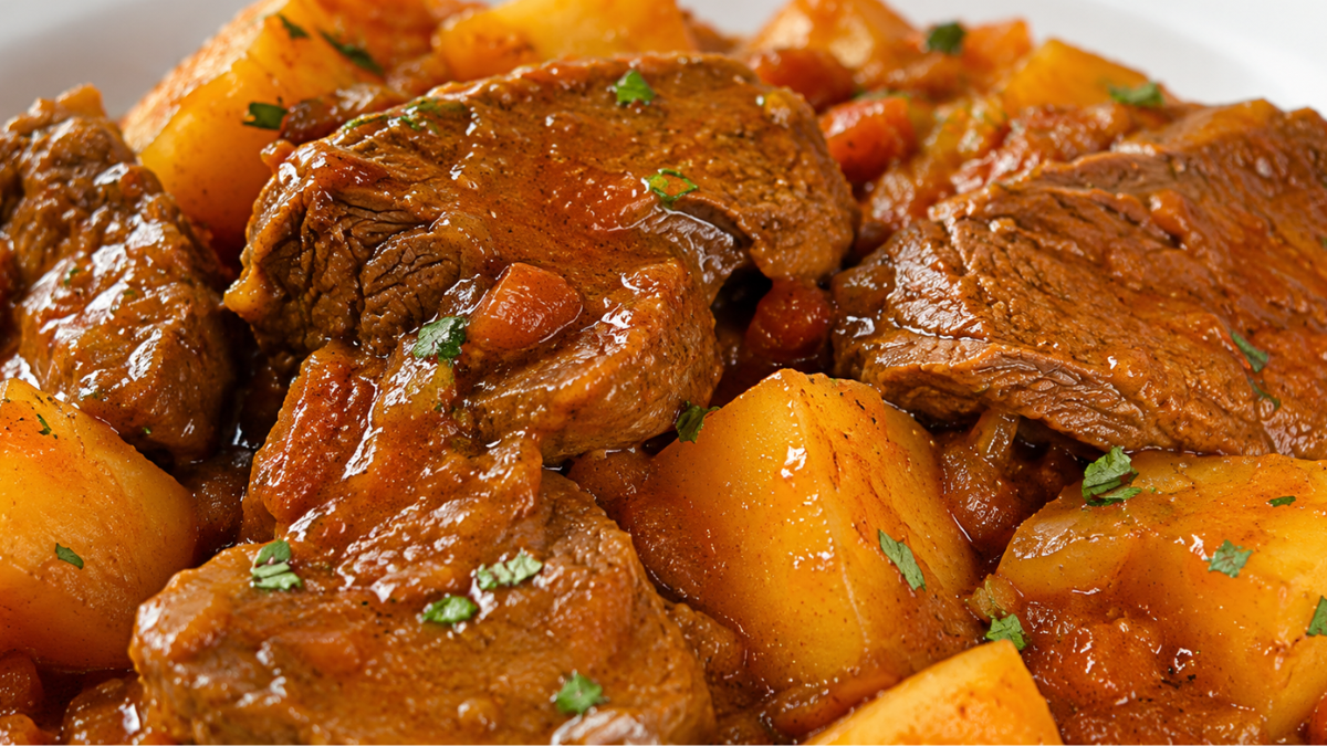 close-up de um prato com fraldinha fatiada em cubos grandes, cozida na panela de pressão, imersa em um molho marrom concentrado com rodelas de cebolas cozidas. O prato está servido com arroz branco e polenta.