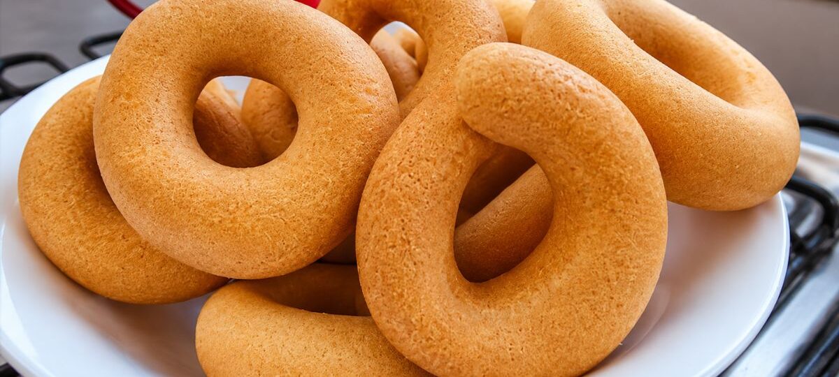 Cesta branca cheia de rosquinhas de polvilho doce douradas e assadas, com uma xícara vermelha ao fundo sobre uma superfície de fogão.