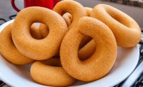 Cesta branca cheia de rosquinhas de polvilho doce douradas e assadas, com uma xícara vermelha ao fundo sobre uma superfície de fogão.