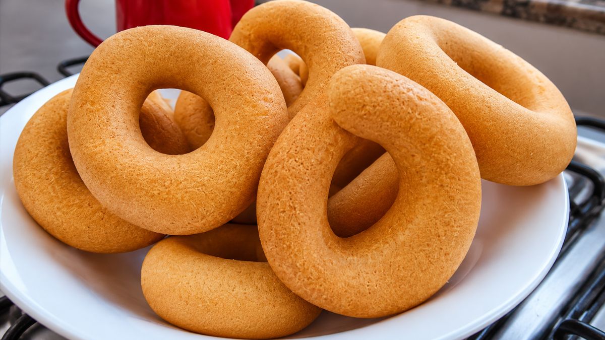 Cesta branca cheia de rosquinhas de polvilho doce douradas e assadas, com uma xícara vermelha ao fundo sobre uma superfície de fogão.