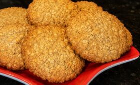 Foto em close de diversos biscoitos caseiros integrais dourados com textura visível de farelo e linhaça dispostos em um prato vermelho sobre uma bancada preta.