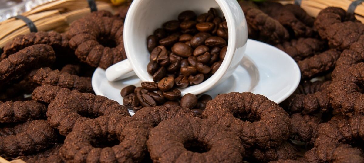 Biscoitos de café caseiros escuros em formato de rosca dispostos em uma cesta de palha rústica com uma xícara branca e grãos de café ao centro.