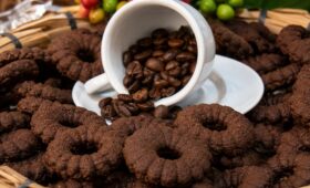 Biscoitos de café caseiros escuros em formato de rosca dispostos em uma cesta de palha rústica com uma xícara branca e grãos de café ao centro.