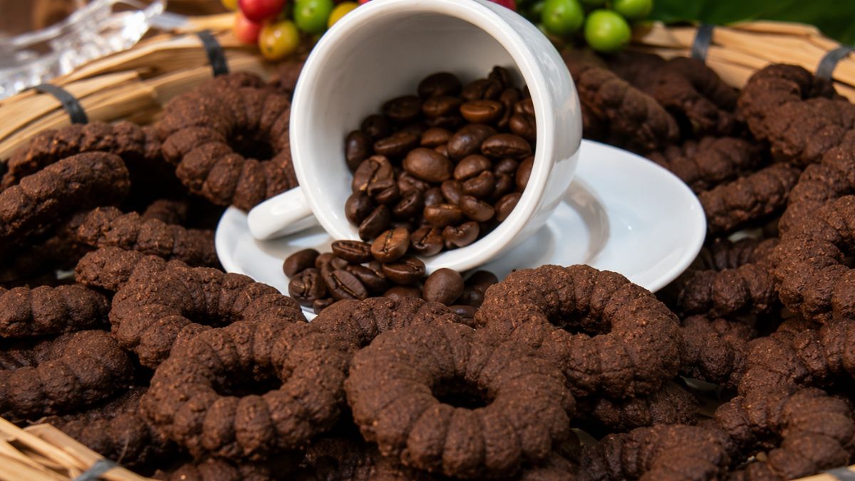 Biscoitos de café caseiros escuros em formato de rosca dispostos em uma cesta de palha rústica com uma xícara branca e grãos de café ao centro.