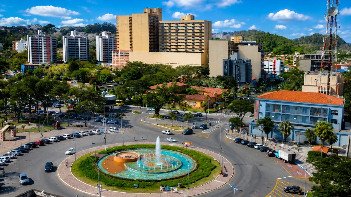 Fotografia aérea de Volta Redonda mostrando a rotatória da Praça Brasil com um chafariz circular ativo, cercada por ruas pavimentadas, carros, edifícios altos e colinas ao fundo.