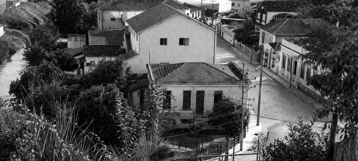 Fotografia aérea em preto e branco mostrando o centro histórico de Mendes, RJ, com casarões antigos, ruas pavimentadas, vegetação e postes.
