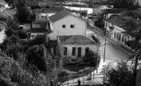 Fotografia aérea em preto e branco mostrando o centro histórico de Mendes, RJ, com casarões antigos, ruas pavimentadas, vegetação e postes.