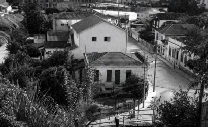 Fotografia aérea em preto e branco mostrando o centro histórico de Mendes, RJ, com casarões antigos, ruas pavimentadas, vegetação e postes.